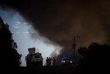 Thick black smoke blankets the sky as two emergency service vehicles respond to a fire.