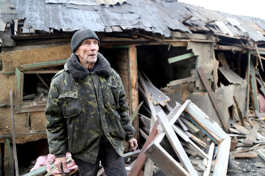 An elderly man in winter clothes, stands in front of a damaged home, among planks of wood and pieces of metal.