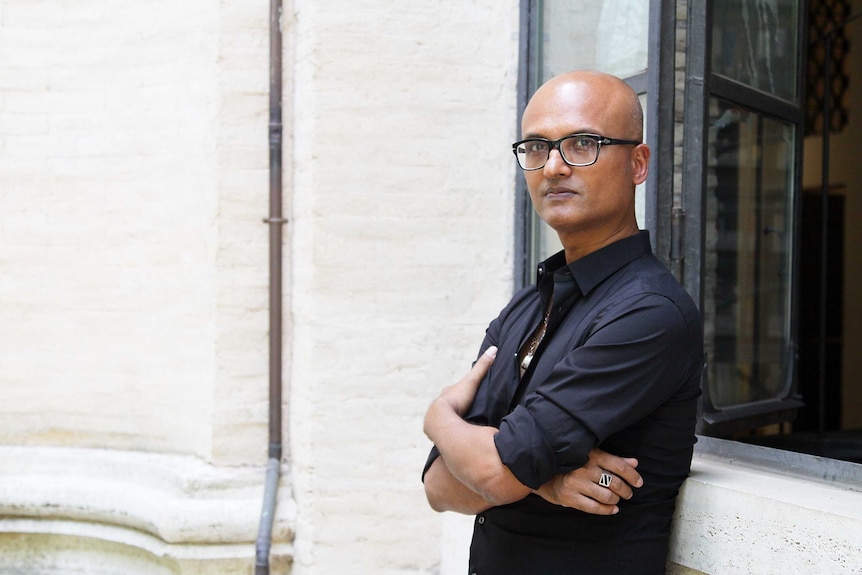 Jeet Thayil stands in front of a window and looks at the camera.