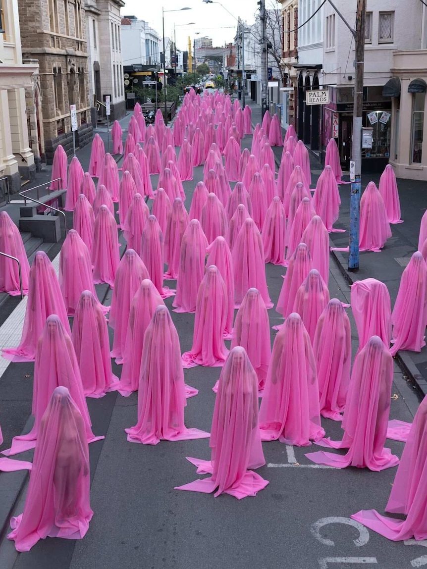 Naked people stand under pink veils.