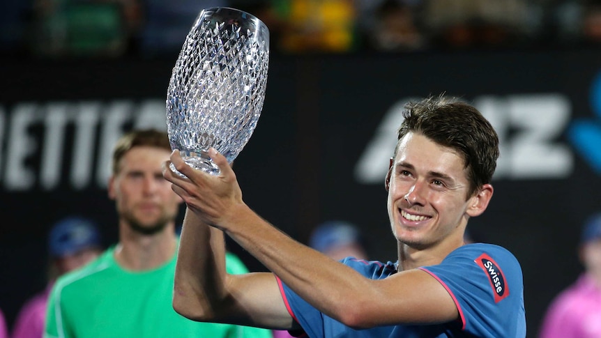 Alex de Minaur grins as he holds a crystal trophy aloft to the left of the frame in both hands while wearing a blue shirt.