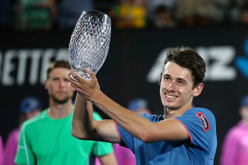 Alex de Minaur grins as he holds a crystal trophy aloft to the left of the frame in both hands while wearing a blue shirt.