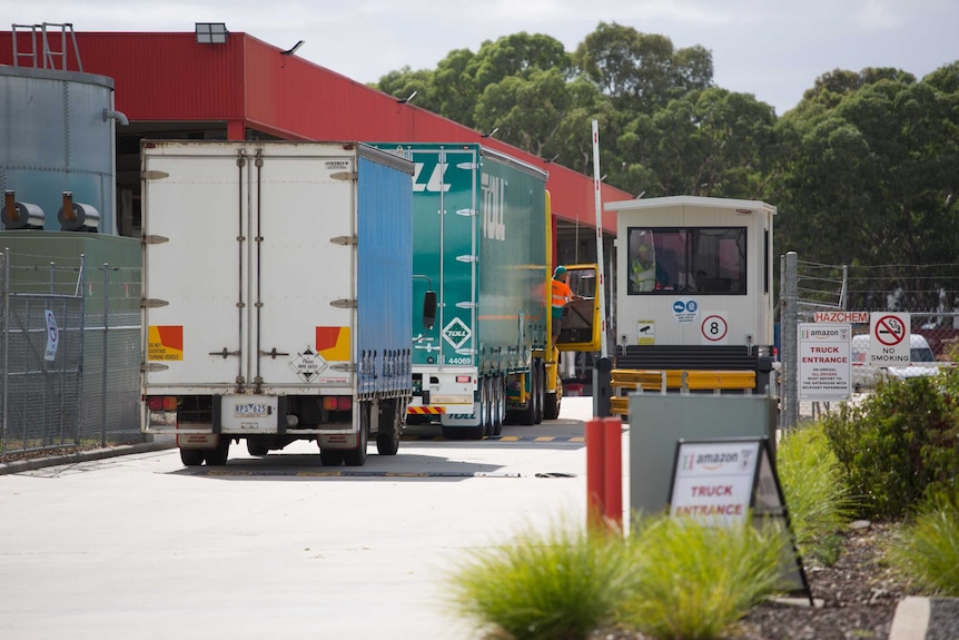 Two trucks stop at the Amazon entry gate, and one driver leans out and talks to security, as a courier prepares to leave.