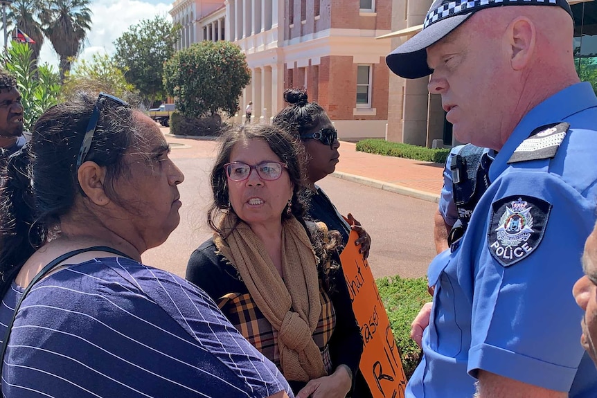 Angry people crowd outside a police station talking to a police officer.