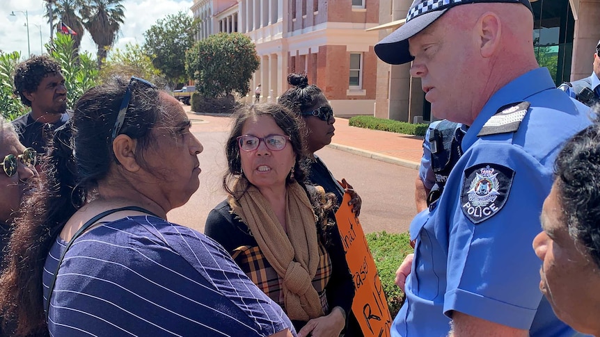 Angry people crowd outside a police station talking to a police officer.