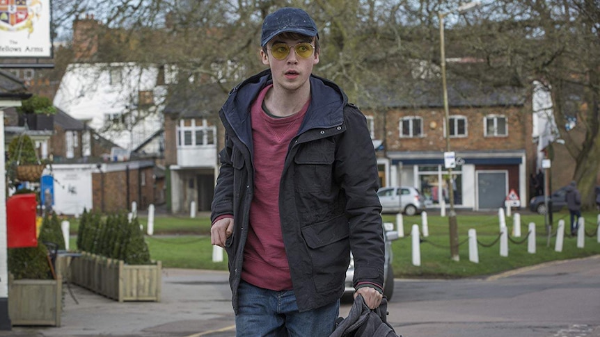 Alex Lawther, playing Kenny in Black Mirror, walks with a bag in hand while wearing sunglasses and a cap.