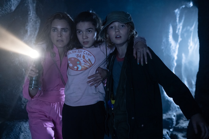 A white woman in a pink jumpsuit and two white boys in casual wear are huddled in a dark cave holding a torch.