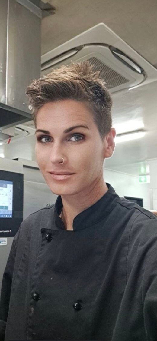 A selfie taken by a brunette with a pixie cut and noise ring. She wears black chefs clothing.
