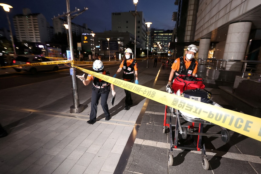 Rescue workers in helmets and masks wheel a stretcher loaded with gear along pavement under a yellow police tape.