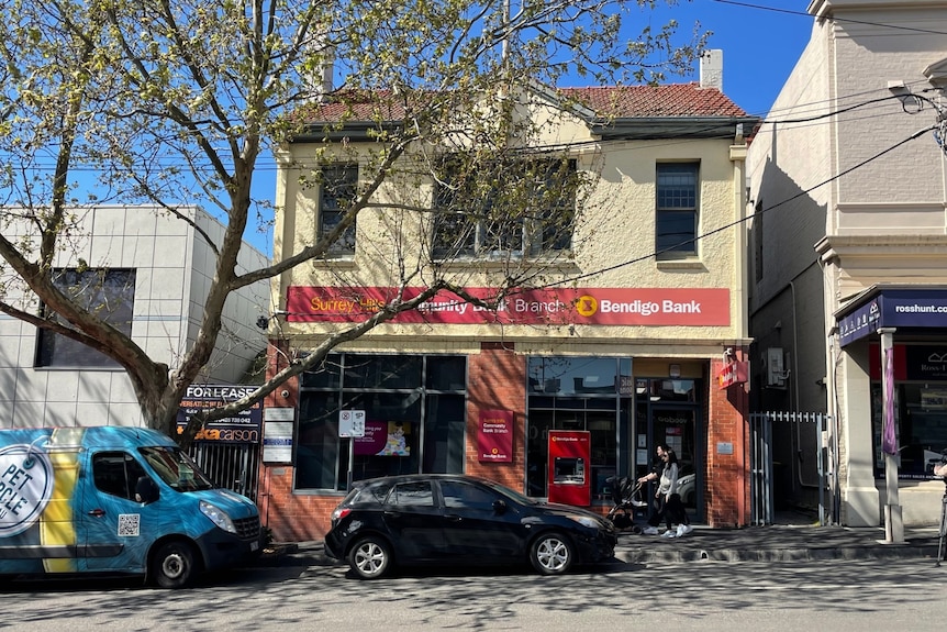 Bendigo Bank at Surrey Hills, there is a black car and a blue van parked on the street in front of it.