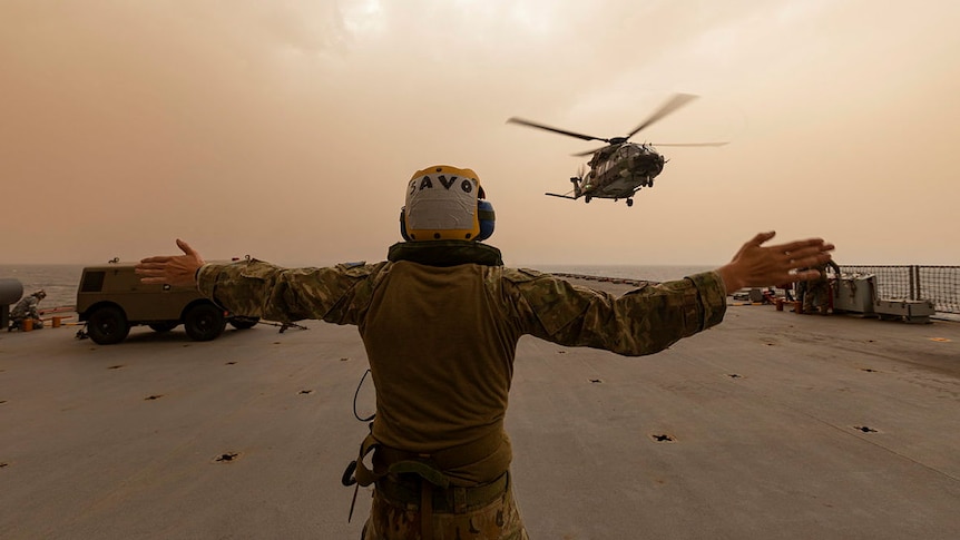 A helicopter approaches a large grey naval base, guided to land by a person wearing uniform with dark smoke filled sky behind