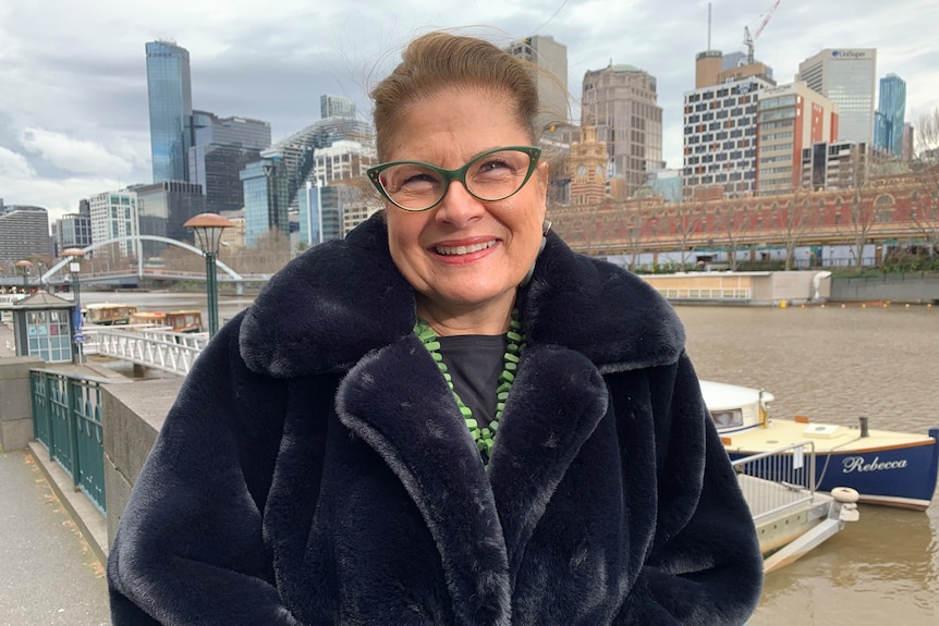 A dark-haired, bespectacled woman in a dark coat stands outside, smiling.