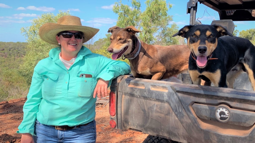 A woman wearing a bright, green shirt, jeans and a hat standing beside two cattle dogs in the back of a farm ute