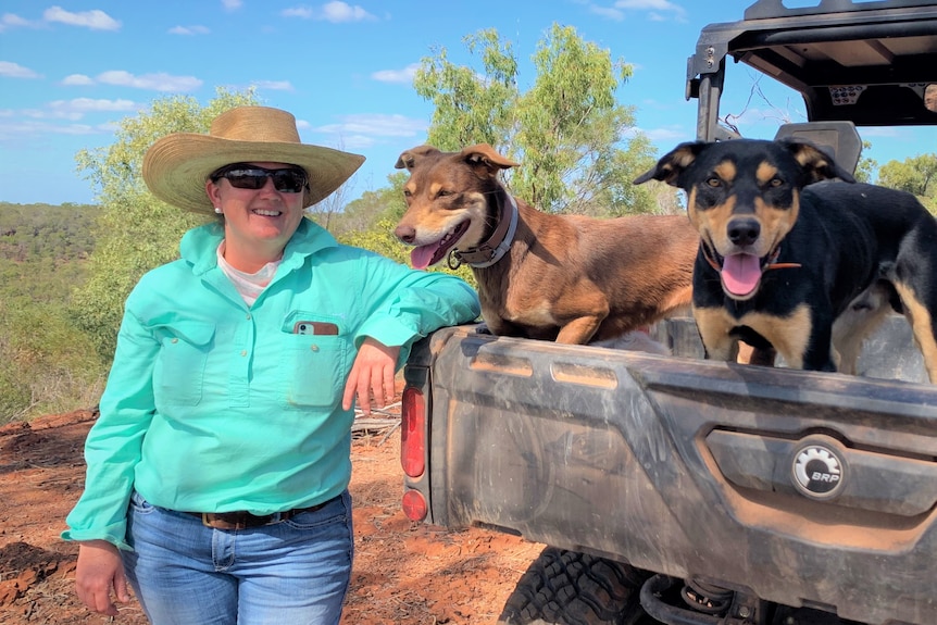 A woman wearing a bright, green shirt, jeans and a hat standing beside two cattle dogs in the back of a farm ute