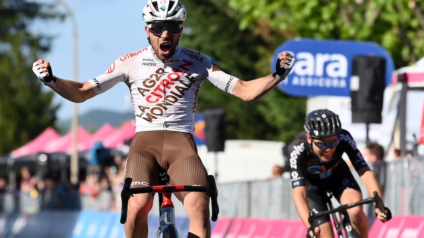 A cyclist celebrates with both arms outstretched as another rider weaves behind him