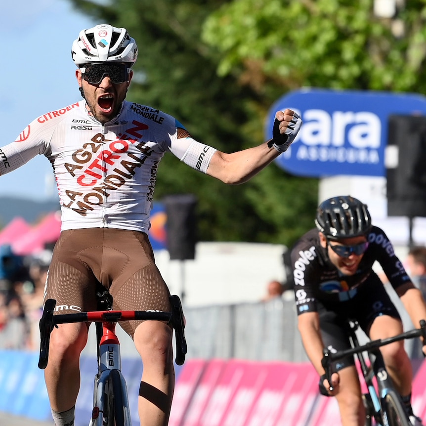 A cyclist celebrates with both arms outstretched as another rider weaves behind him