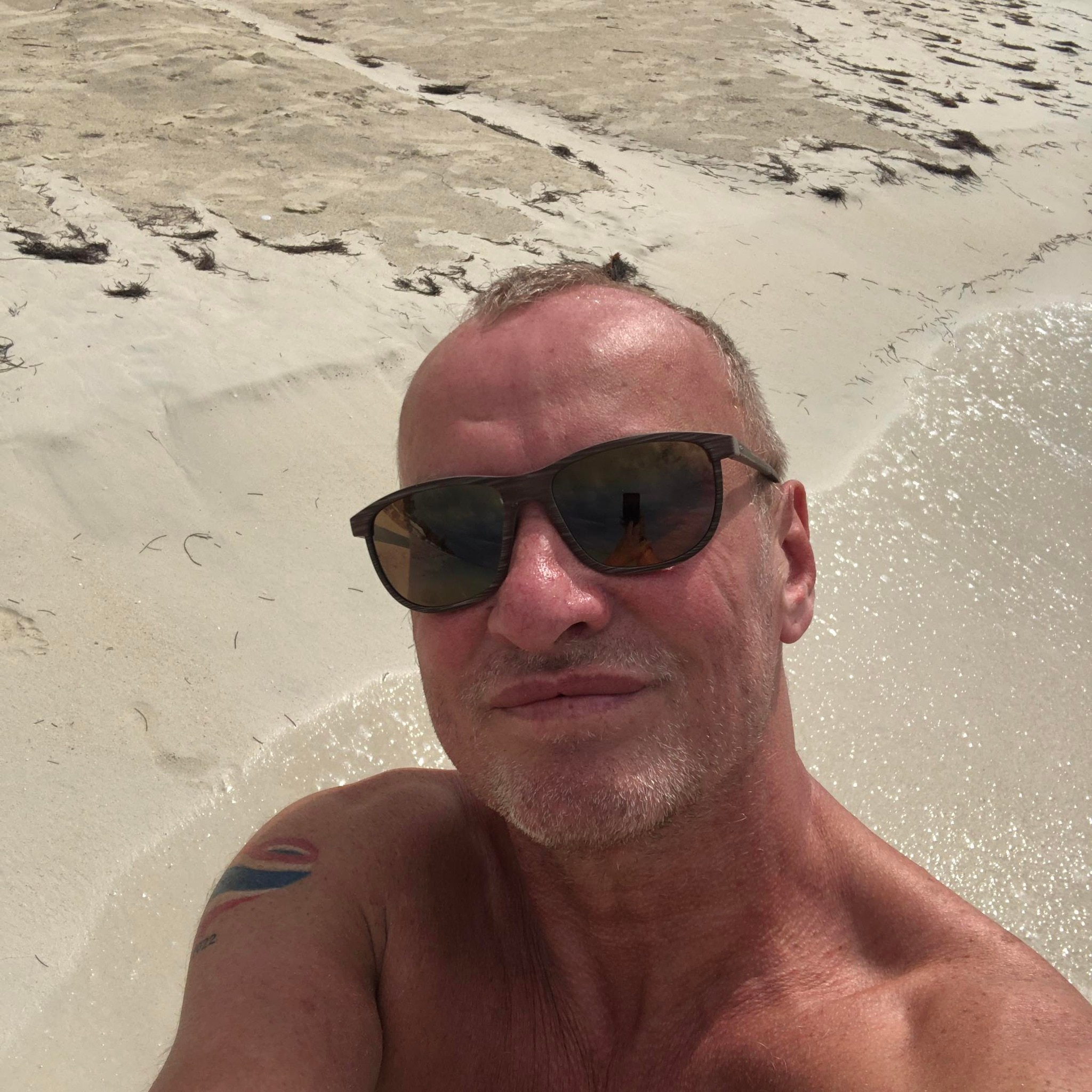 A man wearing black sunglasses at the beach smiles in a selfie.
