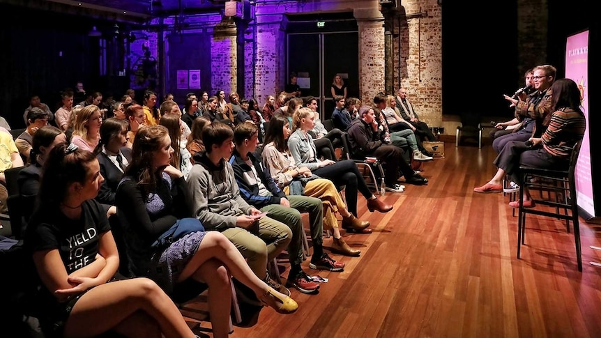 A crowd of young people watch a panel of three people on stools in discussion in a brick auditorium.