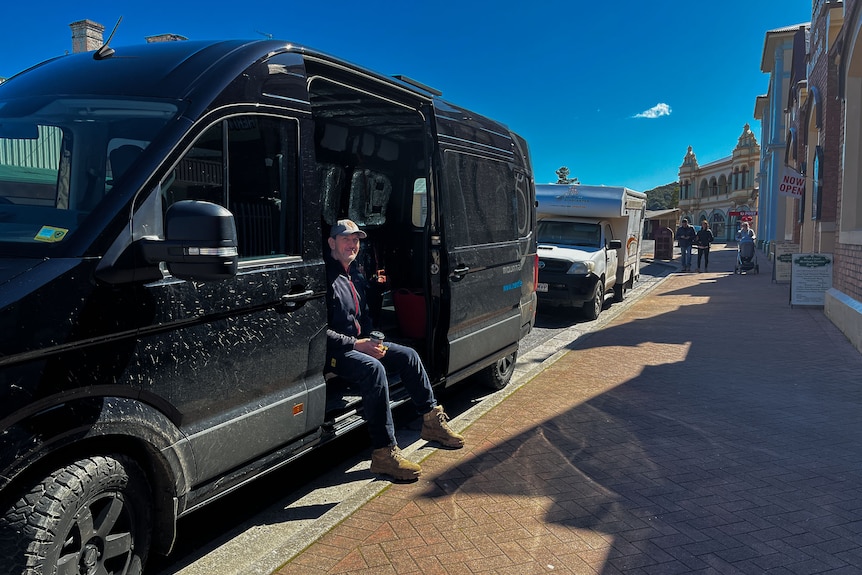 A man holds a takeaway coffee in side door of a large van parked in Zeehan street.