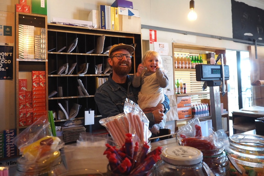 A man in a denim jacket and cap stands behind a shop counter holding a blonde girl.