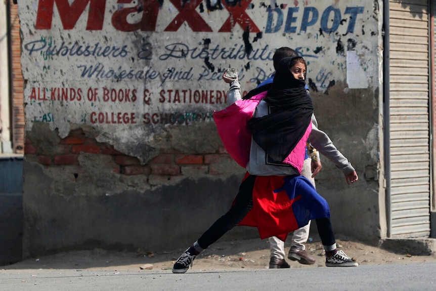 A girl in a black hijab, jeans and sneakers is pictured mid-throw with a rock in her hang in front of a dilapidated building.