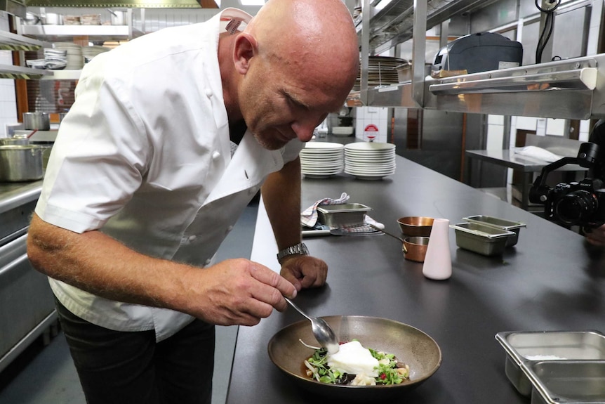 Matt Moran wears white chef's shirt as he bends over to taste toothfish on brown plate in silver kitchen.
