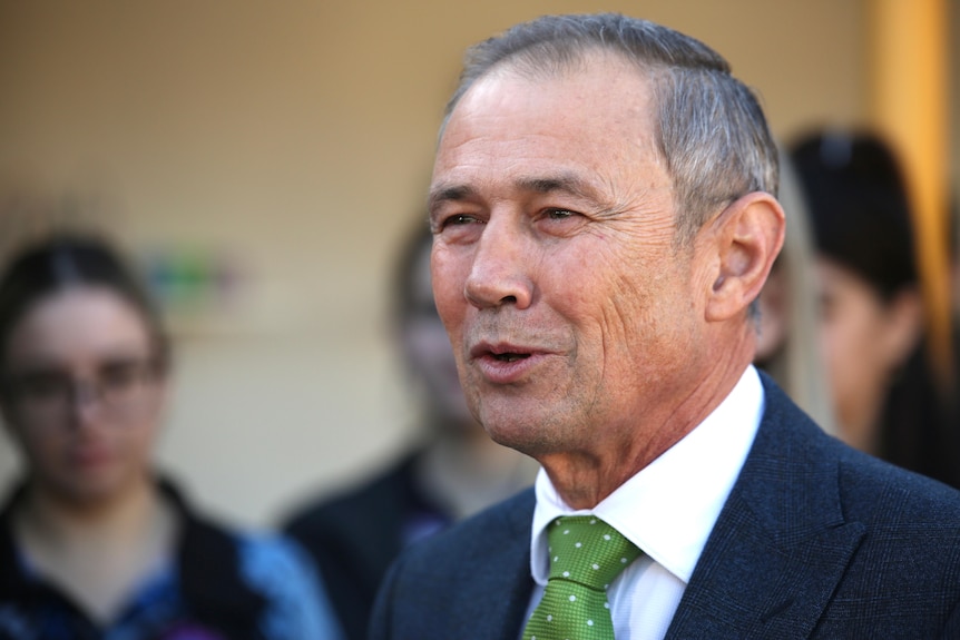A close-up shot of WA Premier Roger Cook smiling wearing a dark suit, white shirt and green tie, with people in the background.