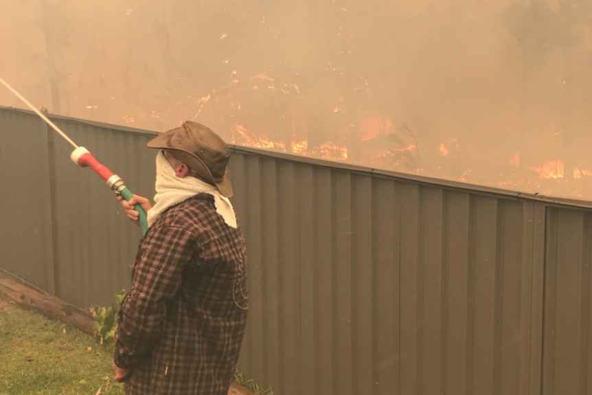 A man with  a towel around his face hoses afire at the fence of his house