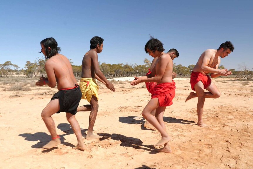 Five Aboriginal boys in red, black and yellow loin cloth dance on yellow dust.