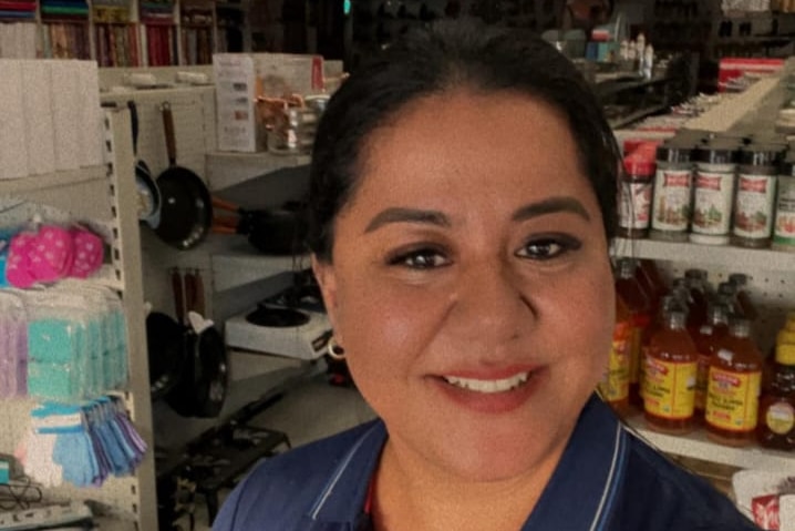 A smiling woman with her hair pulled back takes a selfie inside a small shop, with shelves staked with goods behind her.