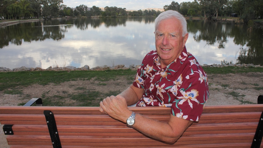 A grey-haired man in a red shirt sitting in front of a lake