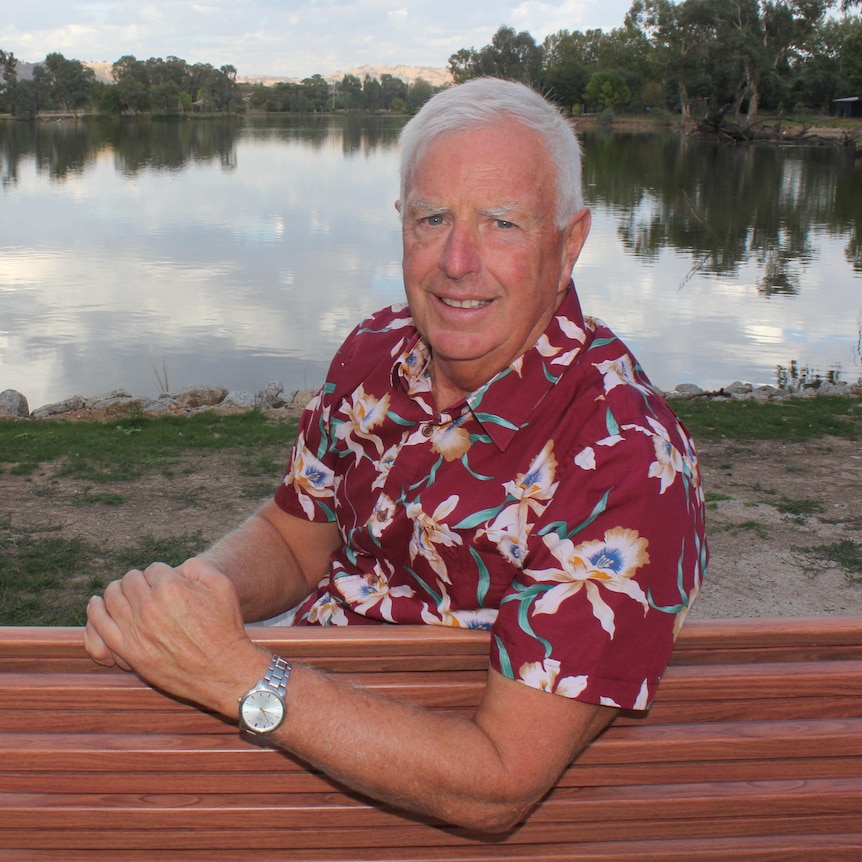 A grey-haired man in a red shirt sitting in front of a lake