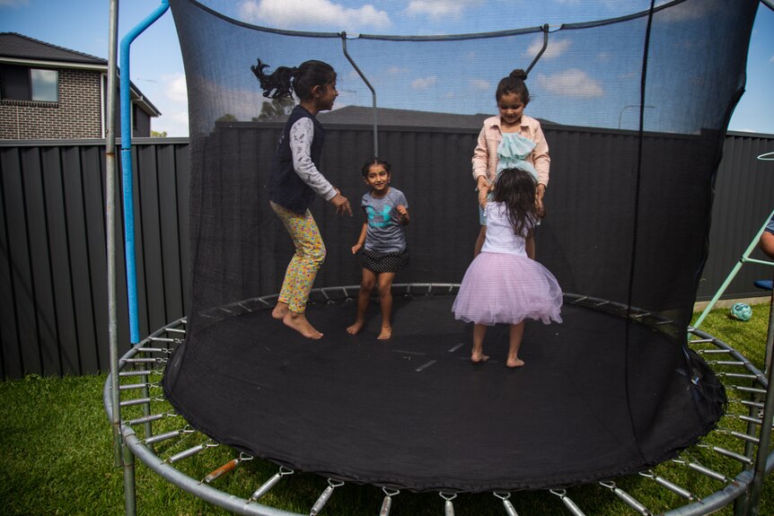 Four children of varying ages bounce on a trampoline in a backyard.