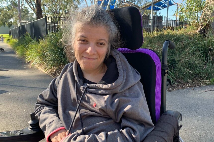 A woman in a grey hoodie sits in her wheelchair in a park