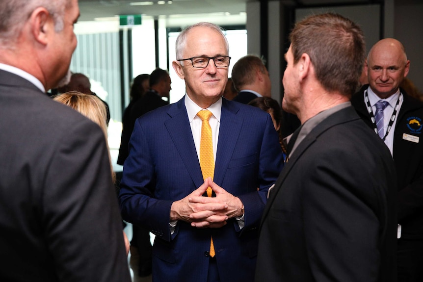 A mid-shot of a smiling Malcolm Turnbull, wearing a blue suit and yellow tie, listening to a man talking to him.