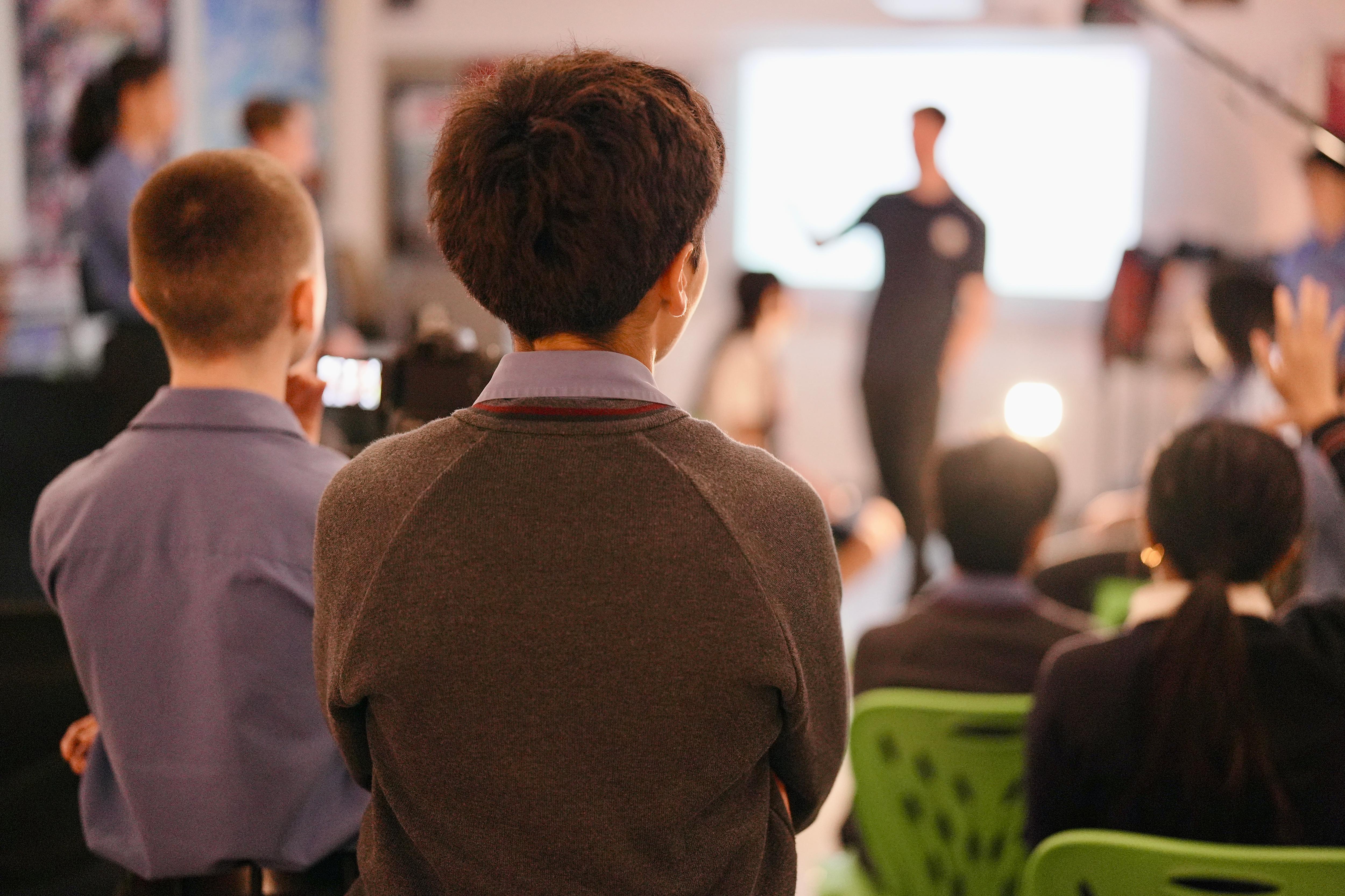 Students stand around listening to a lesson from the teacher in a classroom, only back of heads are visible.
