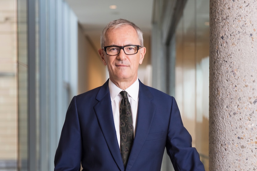 An older man with grey hair and glasses, wearing a suit, looks into the camera, one hand in his pocket