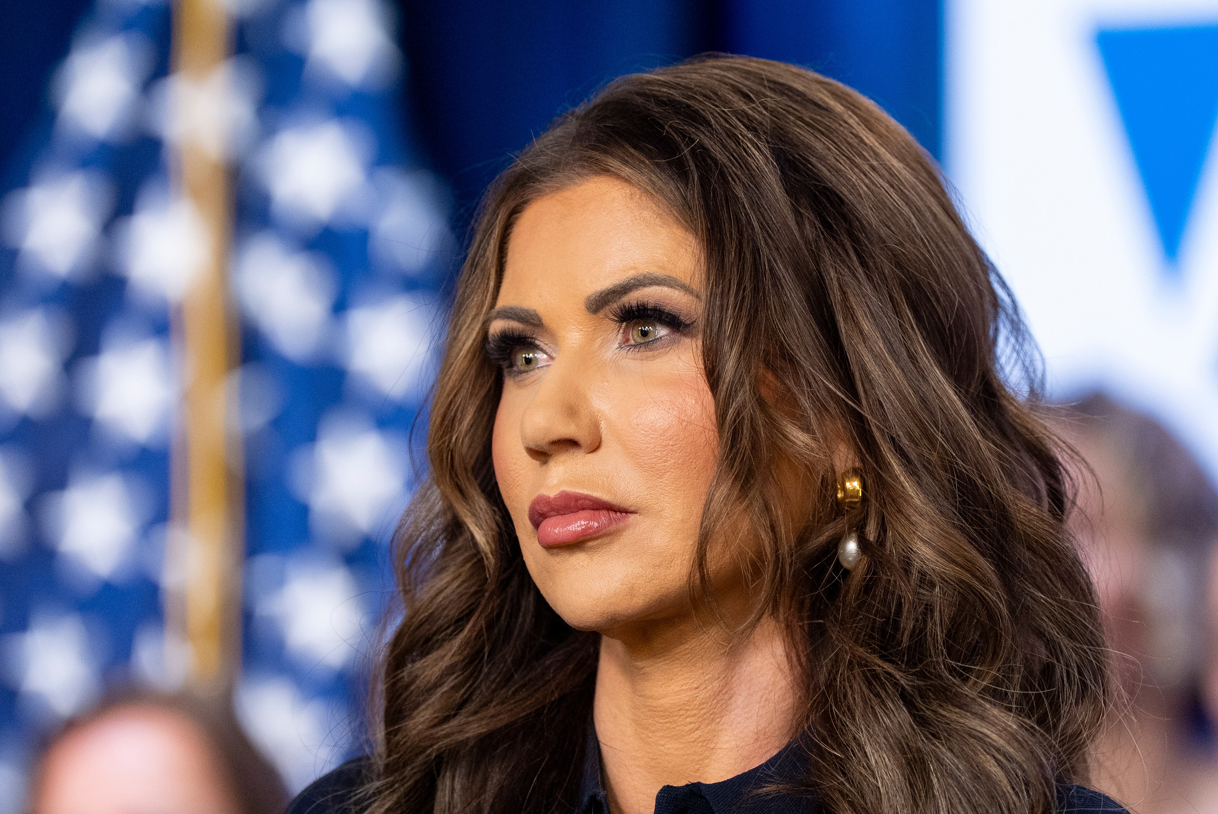 Kristi Noem standing emotionless in a black top in front of a blurred backdrop of a US flag