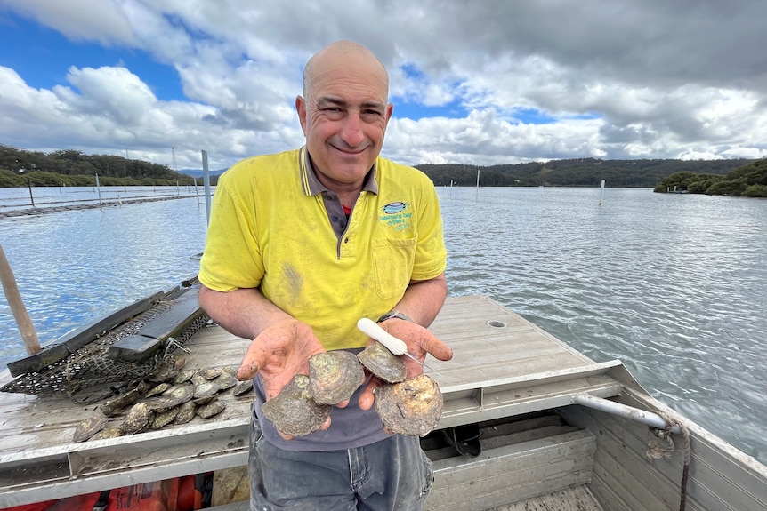 A man stands on a boat holding oysters and a knife. 