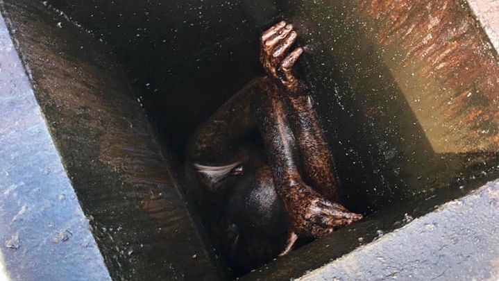 Seen from above, a man is covered in grease and oil, stuck in a metal vent.