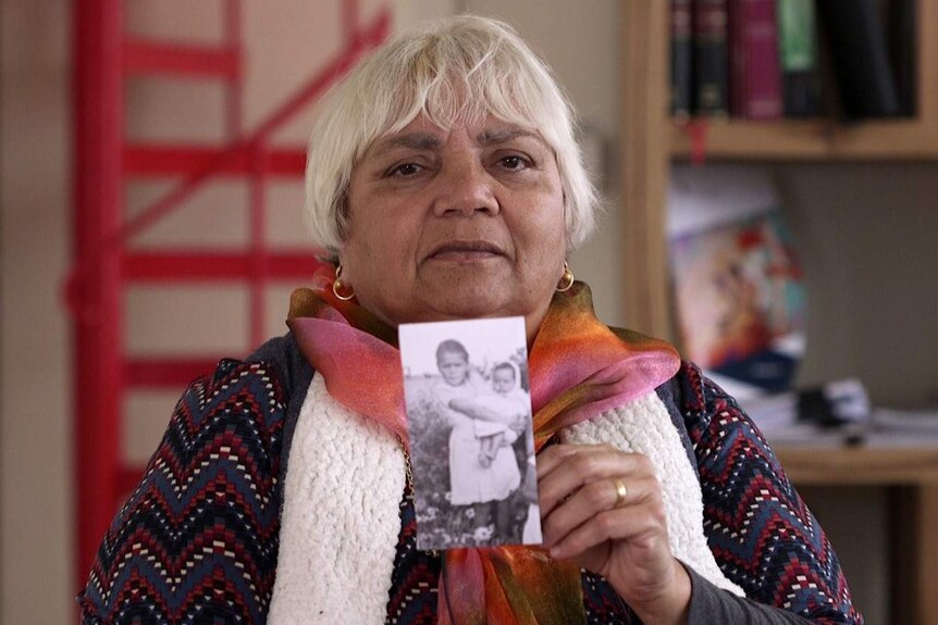 Noongar woman Dallas Phillips holds a photograph of herself as a baby in the arms of her sister standing in front of bushes.
