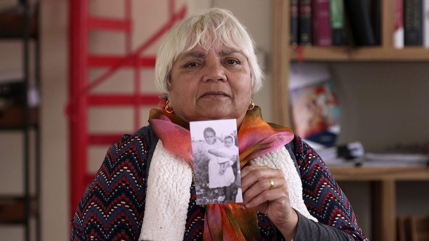 Noongar woman Dallas Phillips holds a photograph of herself as a baby in the arms of her sister standing in front of bushes.