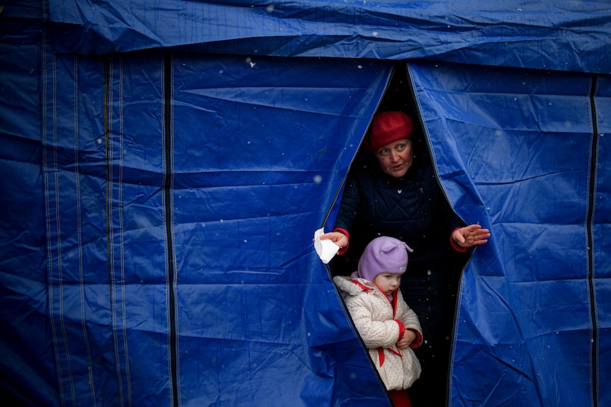 A woman peers through a blue tarpaulin tent with a small child standing in front of her.
