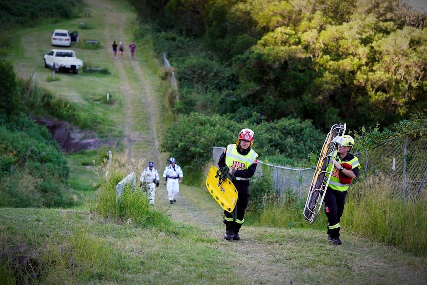 Two rescuers carrying stretchers walk toward the camera down a long grassed area. Behind them are police rescuers also walking