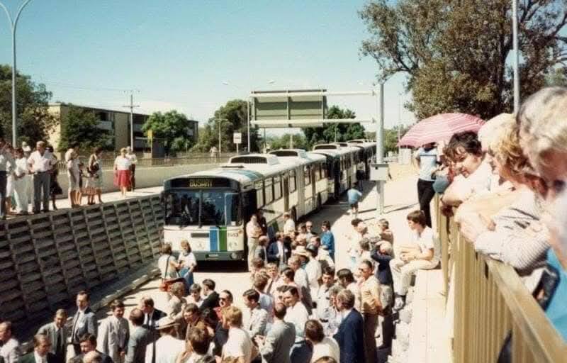 old colour image of a bus on a concrete track with crowds milling infront of it and looking down from above to teh right