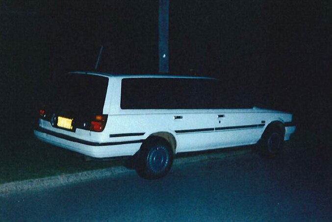 A side-on shot of a white Toyota Camry dating from the 1990s parked on the edge of a grass verge at night.