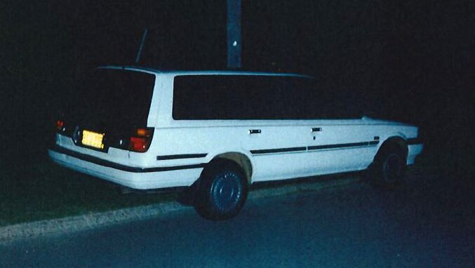 A side-on shot of a white Toyota Camry dating from the 1990s parked on the edge of a grass verge at night.