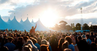 A crowd of people wave their hands in the air in front of tents at sunset.