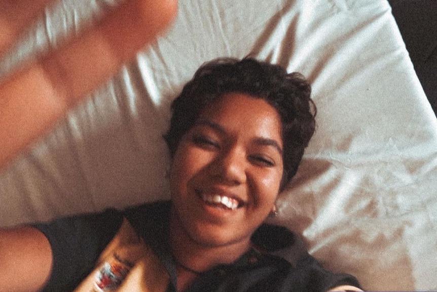 A young woman is seen laying on a bed in a midshot, with one hand stretched towards camera. She smiles.