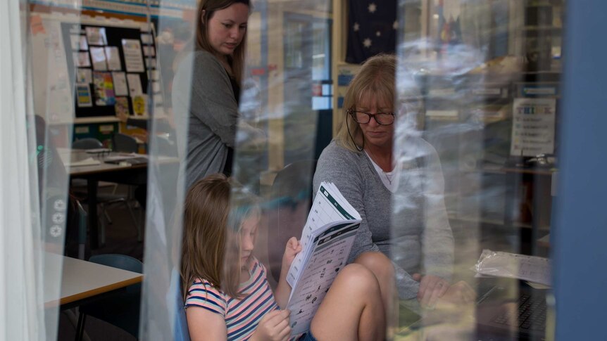 Seen through the fly flaps on a doorway, teacher Anita Harding looks on as a teacher's aide works with a student on reading.